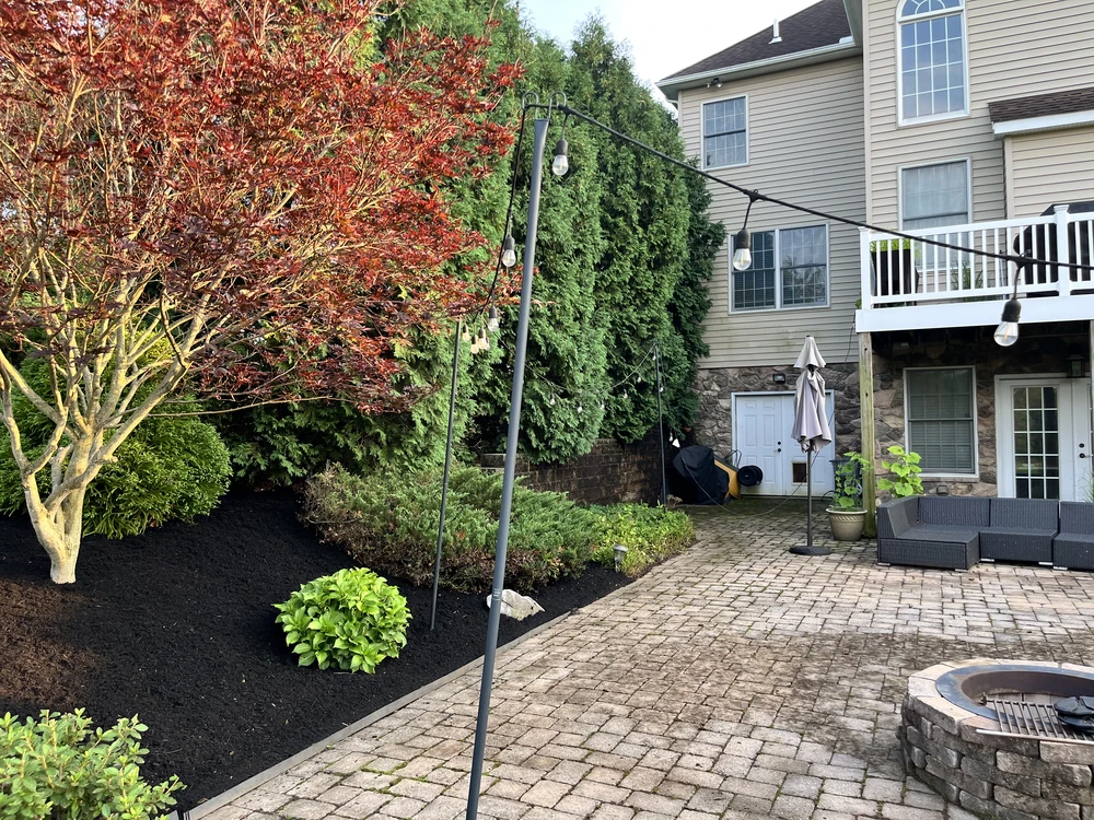 A backyard paver patio with string lights hung on tall black poles, bordered by fresh black mulch, a red-leafed tree, and a stone fire pit, with a multi-story home and dark outdoor furniture in the background.