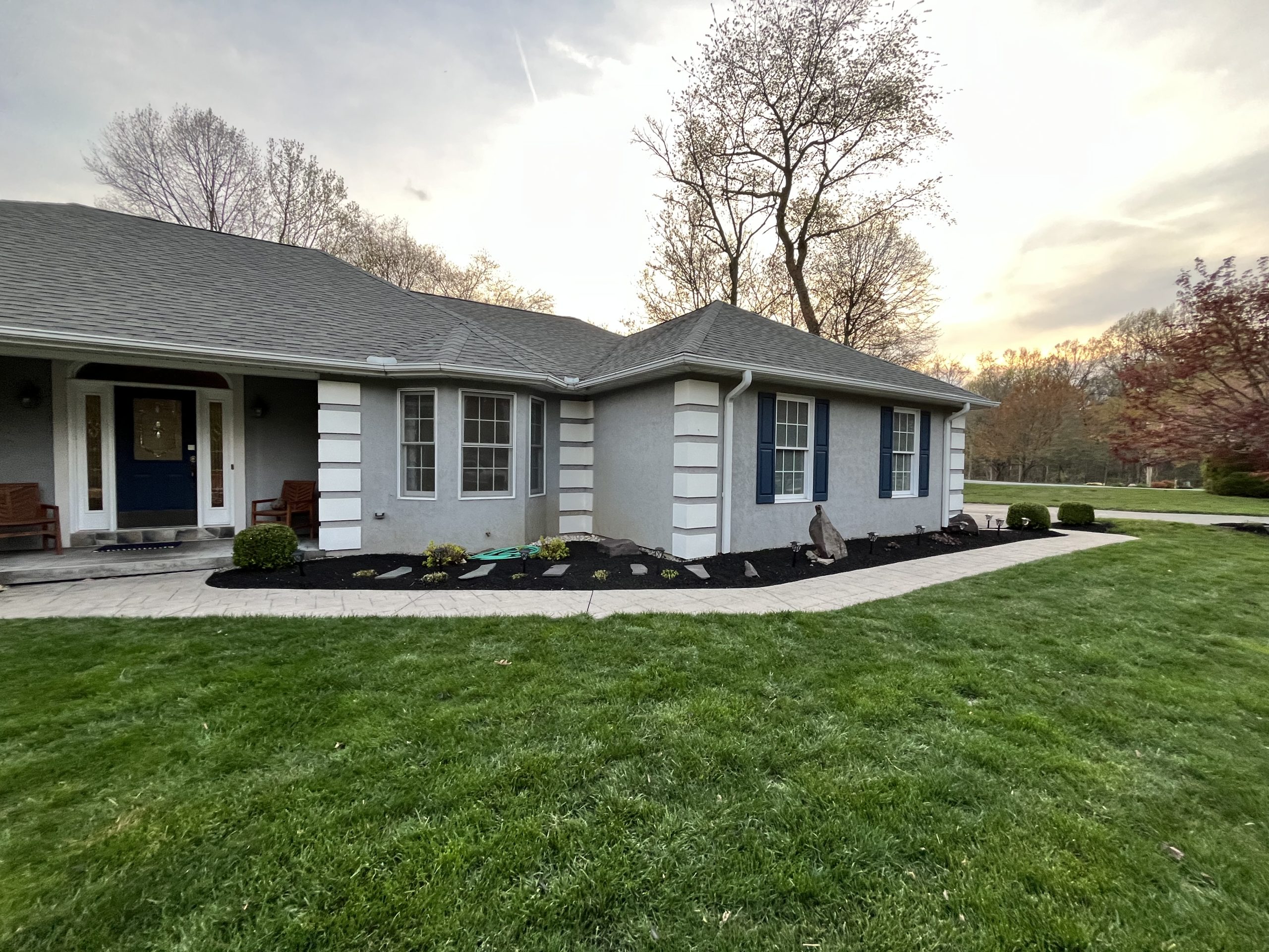 - Landscaping frames a gray home with curved black mulch beds, small shrubs, a pale walkway and a wide green front lawn.