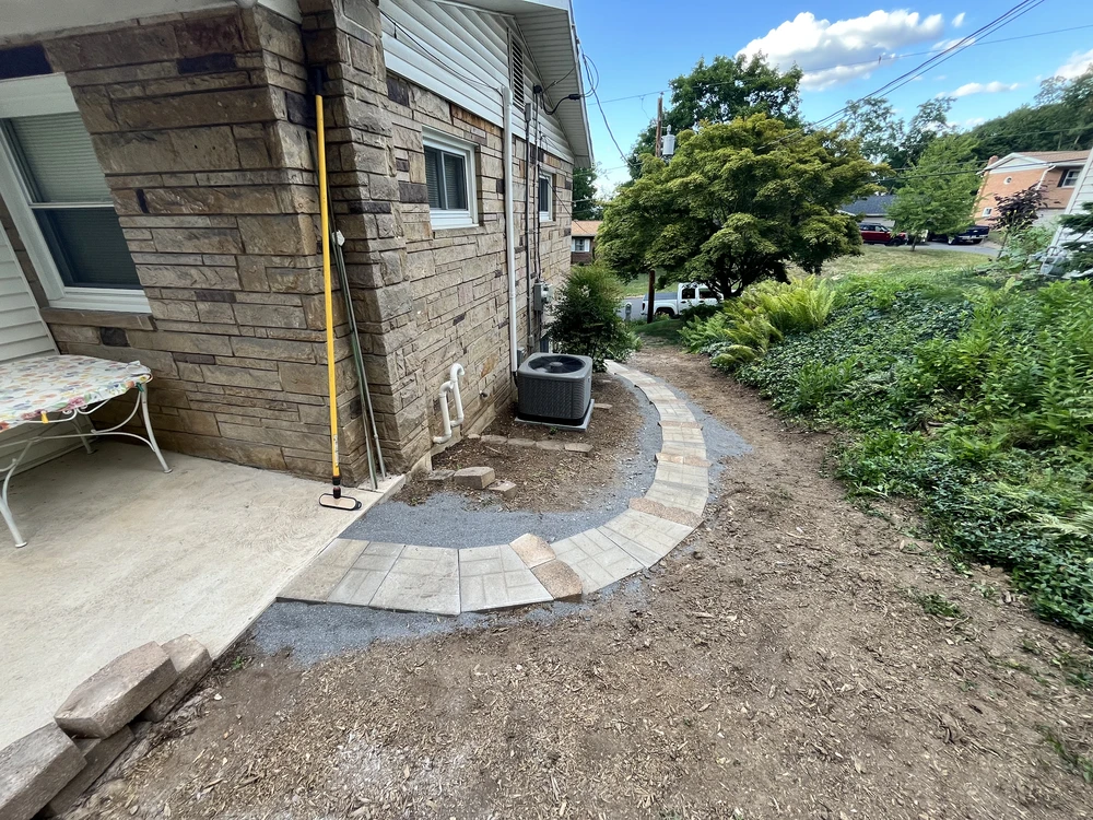 Curved gray and tan paver surface beside a stone home with exposed gravel base and soil on a sloped yard.