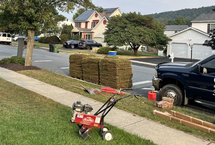 Stacks of rolled sod, a red tiller, hand tools, and a work truck beside a residential sidewalk before installation.
