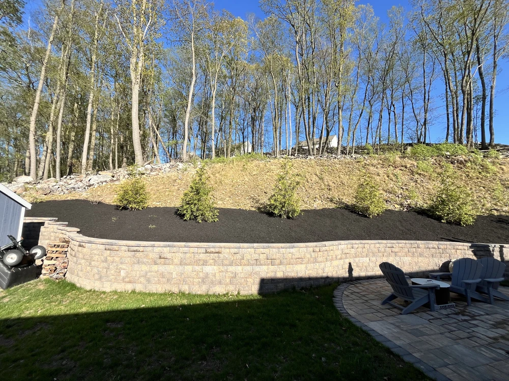 Fresh black mulch and young shrubs above a block retaining wall beside a small paver seating area on a wooded slope.